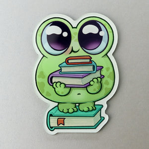A "Read-A-Bit" Acrylic Magnet of a cute cartoon frog with large eyes hugging a stack of colorful books, standing on top of another book from the Thimblestump Hollow series, made by Bindlewood Shop.
