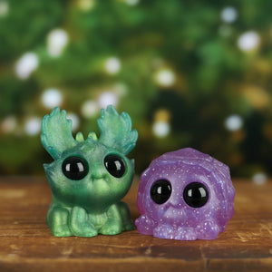 Two adorable, Chris Ryniak Holiday Cheer Sets with large, expressive eyes sitting on a wooden surface against a blurred background of greenery and white flowers.