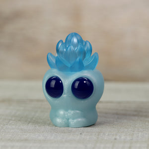 A small, whimsical toy sculpted with a translucent blue flame-like top and large, glossy black eyes, resembling a cute ghost, set against a neutral wooden background. - Ice Frost Archie by Chris Ryniak