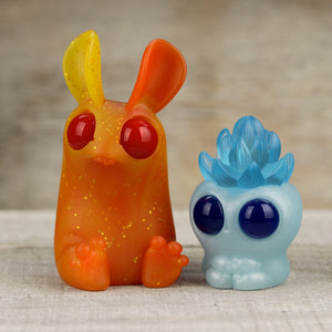 Two colorful, whimsical resin figures: one resembles an orange rabbit with a yellow ear and red eyes, and the other is a blue creature with large eyes and a floral-like top from the Fire & Ice Set by Chris Ryniak.