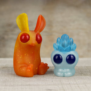 Two colorful, whimsical resin figures from the Chris Ryniak Fire & Ice set—a sparkly orange rabbit-like figure with red eyes, and a blue, crystal-textured creature with large blue eyes—on a wooden.