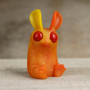 A small, vibrant orange resin figure of a cartoon-like rabbit with oversized red eyes and prominent ears, one of which is bent, designed by Amanda Louise Spayd and set against a neutral backdrop by Bindlewood Shop's Sparkle Flame Peppercorn.