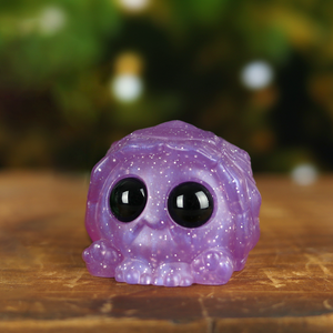 A sparkly purple Sugar Plum Turtball, designed by Chris Ryniak, with large, glossy black eyes sitting on a wooden surface with a blurred green background.