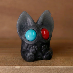 A small, dark, cat-like figurine with large, round blue and red eyes. The figurine has pointy ears and detailed paws, sitting on a wooden surface. Crafted in the style of Chris Ryniak's 3D Matinee Set resin figures, it has a speckled, starry texture reminiscent of Smidgen and Dashi characters.