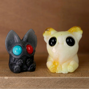 A photo of two small, cute resin figures from the 3D Matinee Set by Chris Ryniak, set on a wooden surface. The figurine on the left is a dark-colored cat with large, mismatched eyes—one blue and one red. The figurine on the right is Smidgen, a light-colored creature with large round black eyes and small ears.