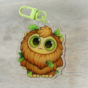 A cute, cartoonish keychain designed by Amanda Spayd, featuring a fluffy, brown creature with big green eyes and leafy accents from Thimblestump Hollow, set against a wood "Bigfoot" Acrylic Charm from Bindlewood Shop.