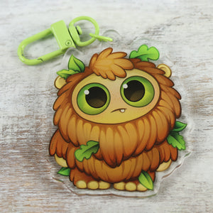 A cute, cartoon-style keychain featuring a whimsical, fluffy, brown creature with big green eyes and leafy accents from the Bindlewood Shop collection.