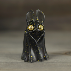 The Black & Gold Belfry by Bindlewood Shop is a smoke-black resin figure with gold glitter, large gold eyes, upright ears, and a cloak-like garment, designed by Amanda Louise Spayd and photographed against a blurred neutral background.