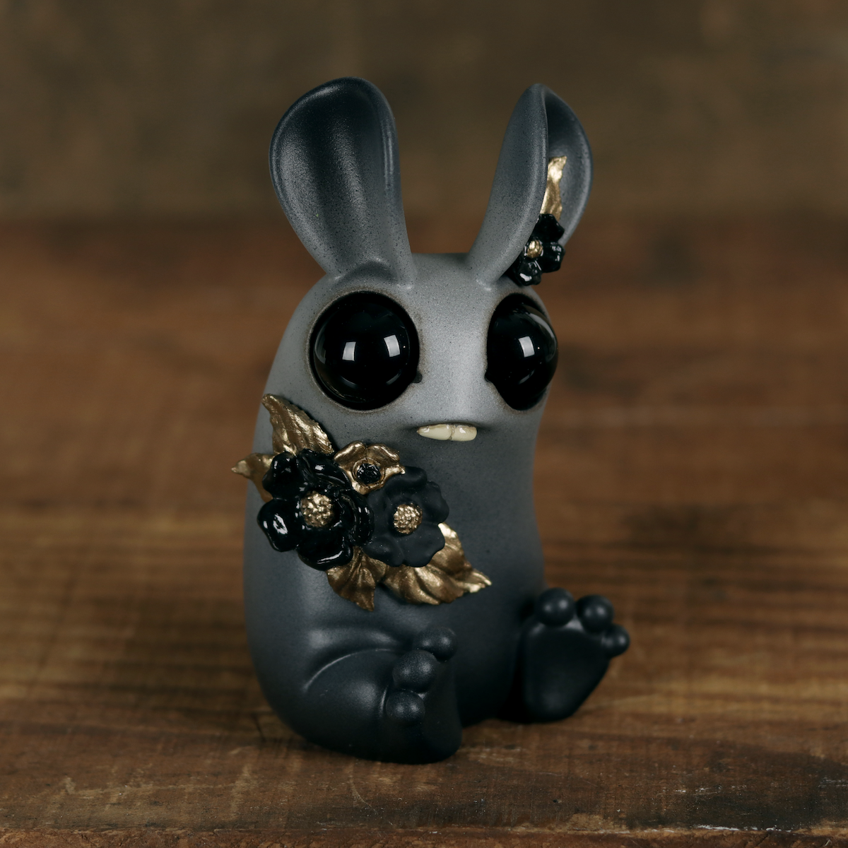 The Amanda L Spayd Black & Gold Peppercorn resin figurine is a small, dark gray, rabbit-like creature featuring large black eyes, tiny fangs, and hand-painted gold-accented black flowers adorning its body. Set on a wooden surface against a softly blurred brown background, this charming piece embodies an enchanting and whimsical allure characteristic of Amanda L Spayd’s unique style.