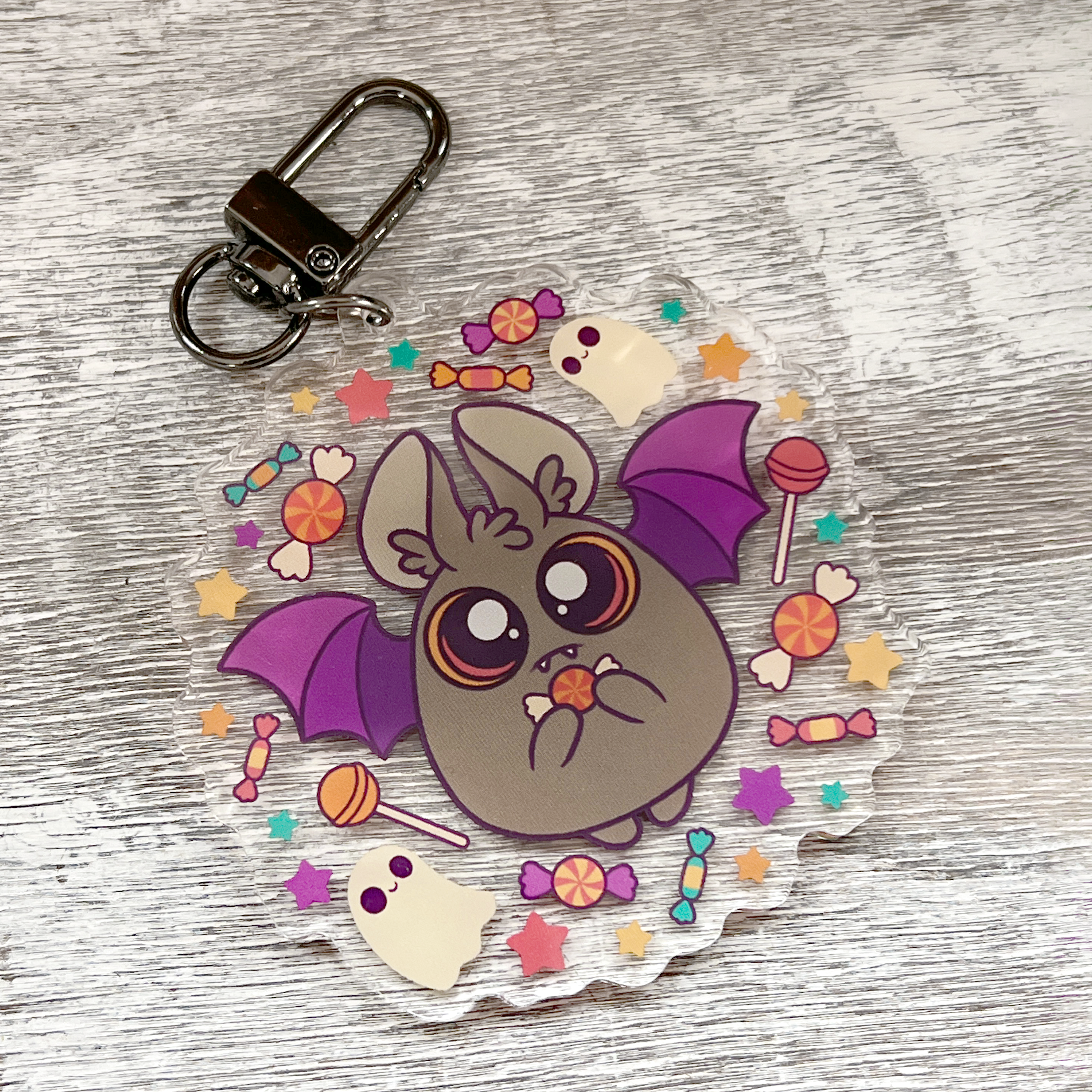 The CandyBat Keychain from Bindlewood Shop is a round acrylic charm featuring a cute cartoon bat with big eyes and purple wings holding candy. Inspired by Amanda Louise Spayd, the illustration shows the bat surrounded by colorful candies, stars, and tiny white ghosts on a light wood background.