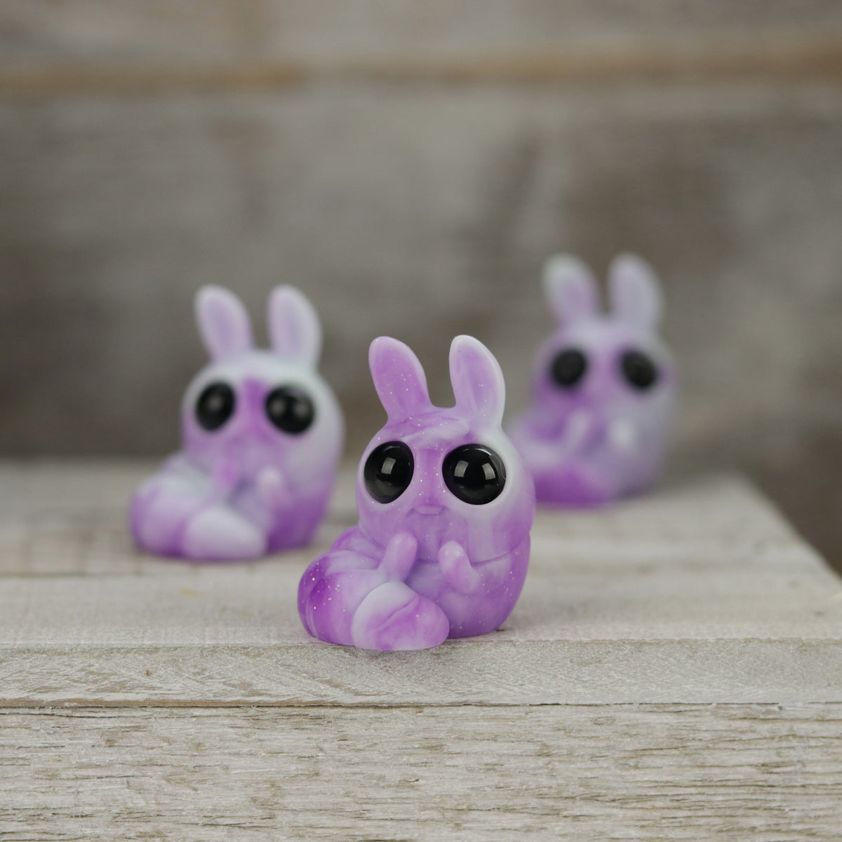 Three small, purple Plasma Storm Chenille figures from Bindlewood Shop, with big black eyes and a glossy, marbled finish, are arranged on wood. Their cute design is inspired by Amanda Louise Spayd and evokes a whimsical charm.