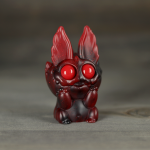 The Midnight Cranberry Nettlepup by Bindlewood Shop is a small red and black figurine with large round eyes and flame-like ears, sitting on a gray surface with glittery red swirl accents and an adorably surprised expression.