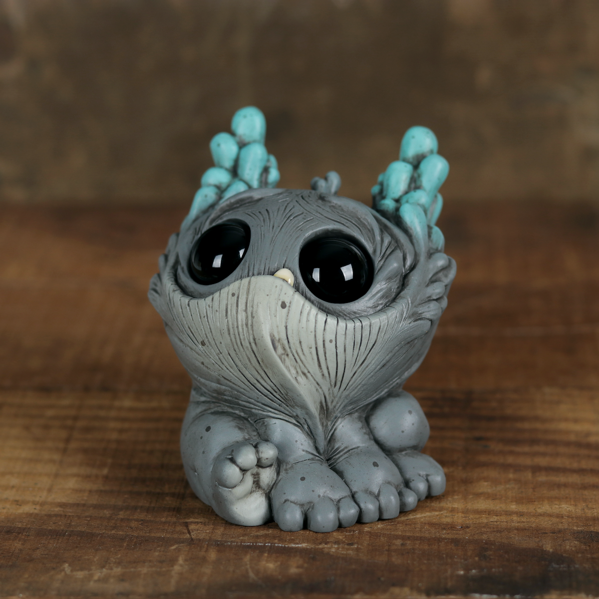 A small, gray Cave Glow Dewlap figurine—crafted by Chris Ryniak in his signature whimsical style—sits on a wooden surface against a brown background. Featuring large black eyes, textured fur, big feet, and turquoise bumps on its head, this expertly hand-painted solid resin collectible is sure to captivate fans of unique art toys.