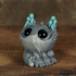 A small, gray Cave Glow Dewlap figurine—crafted by Chris Ryniak in his signature whimsical style—sits on a wooden surface against a brown background. Featuring large black eyes, textured fur, big feet, and turquoise bumps on its head, this expertly hand-painted solid resin collectible is sure to captivate fans of unique art toys.