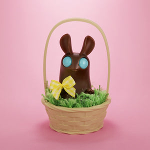 The Chocolate Bunny Basket Peppercorn by Bindlewood Shop features a blue-eyed chocolate bunny with a yellow checked bow, nestled in a small wicker basket with green grass and a printed base, set against a pink background.