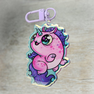 A colorful Flooficorn keychain with a whimsical, Thimblestump Hollow-inspired design, featuring a chubby pink character with a purple mane and a single horn, playfully posed against a wooden background by Bindlewood Shop.
