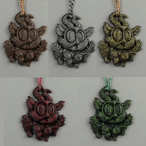 Five Glitter Krampus Ornaments by Bindlewood Shop—textured pendants shaped like horned creatures with big eyes, each in a different metallic hue—are displayed on a gray background with matching cords, sparkling like tiny resin treasures.