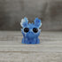 The Sunshower Moose by Bindlewood Shop is a small blue fantasy resin figure with big black eyes, white-tipped antlers, and a star-speckled pattern, shown sitting on a wooden surface against a blurred wooden background.