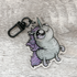 Presenting the Goth Ponycorn Keychain by Bindlewood Shop—a double-sided acrylic charm portraying a cartoon unicorn with a large eye, gray body, and purple mane and tail, lying on its back against a textured gray wooden background—perfect for fans of unique unicorn accessories.