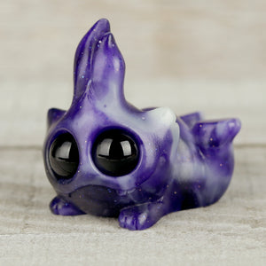 The Mooncloud Bugbite by Chris Ryniak is a small, purple resin figure with a galaxy pattern, large shiny black eyes, and a whimsical aquatic-inspired shape resting on a light wooden surface.