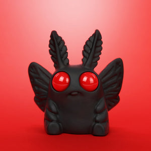 Amanda L Spayd’s Mothman Edison is a small black figurine with moth-like wings, large red eyes, and leaf-shaped antennae. Crafted from opaque resin, it’s shown against a vibrant red background.