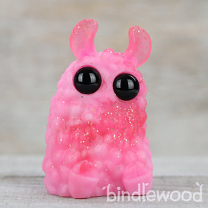 The Raspberry Sparkle Crème PomPom, a small pink textured resin figure with large black eyes, glittery body, and translucent horns, sits on light wood. "Bindlewood Shop" appears with artist Amanda Louise Spayd at the bottom right.