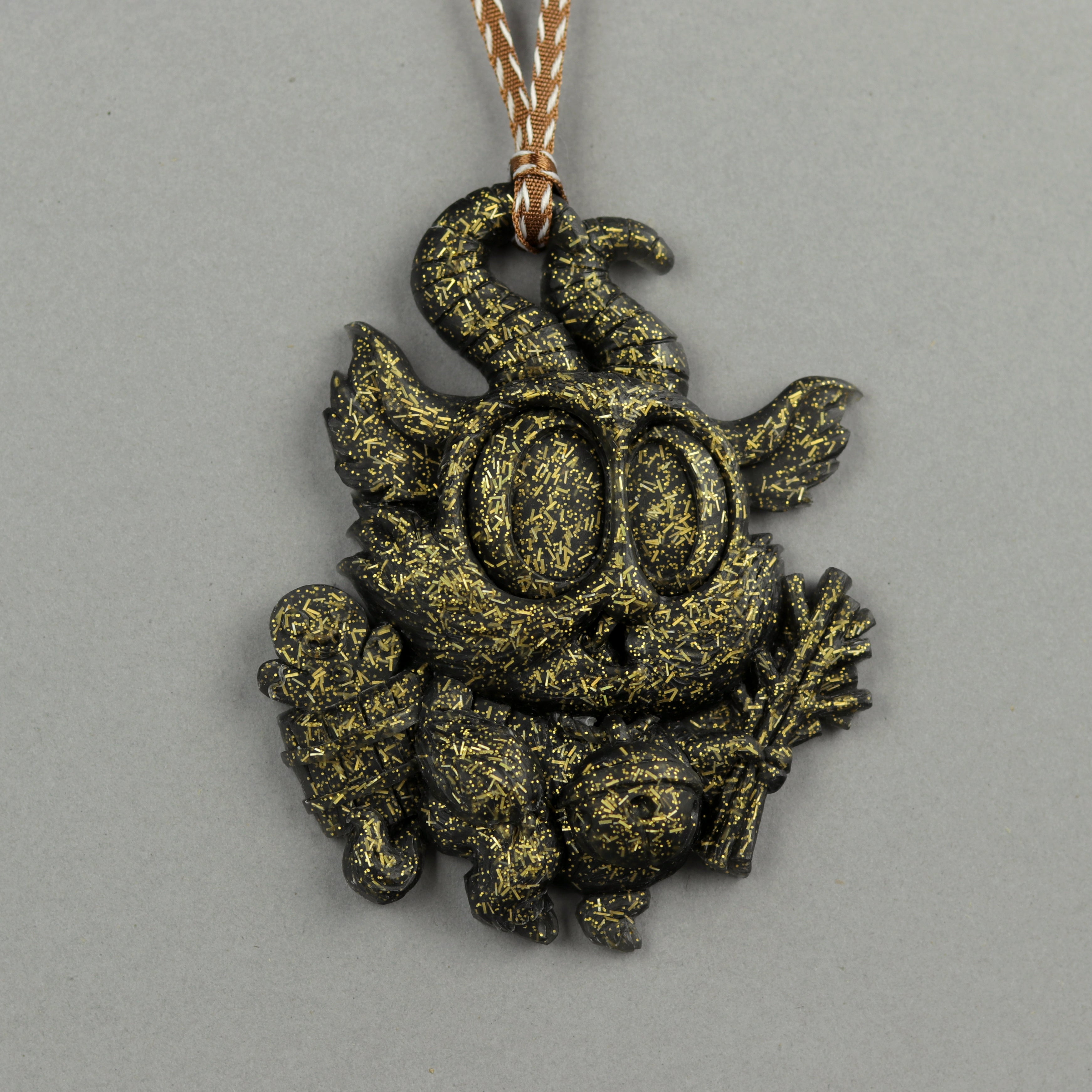 The Glitter Krampus Ornament by Bindlewood Shop features a whimsical creature with large eyes, horns, wings, and twigs. It has a black and gold speckled finish and hangs from a striped cord against a gray background.