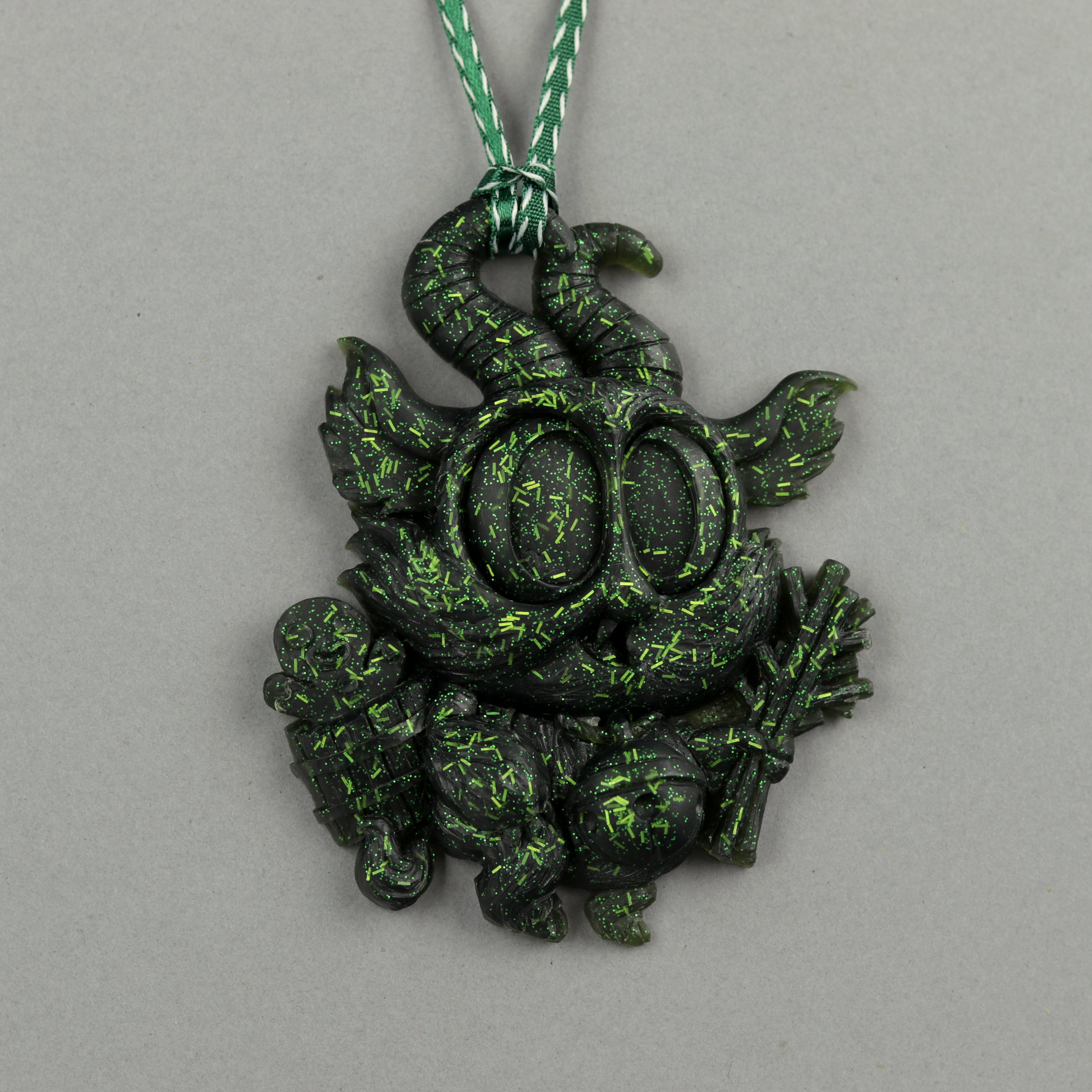 The Bindlewood Shop Glitter Krampus Ornaments feature a horned, big-eyed creature with a staff, sitting cross-legged. Black with green speckles, it hangs from a green and white cord on a gray background.