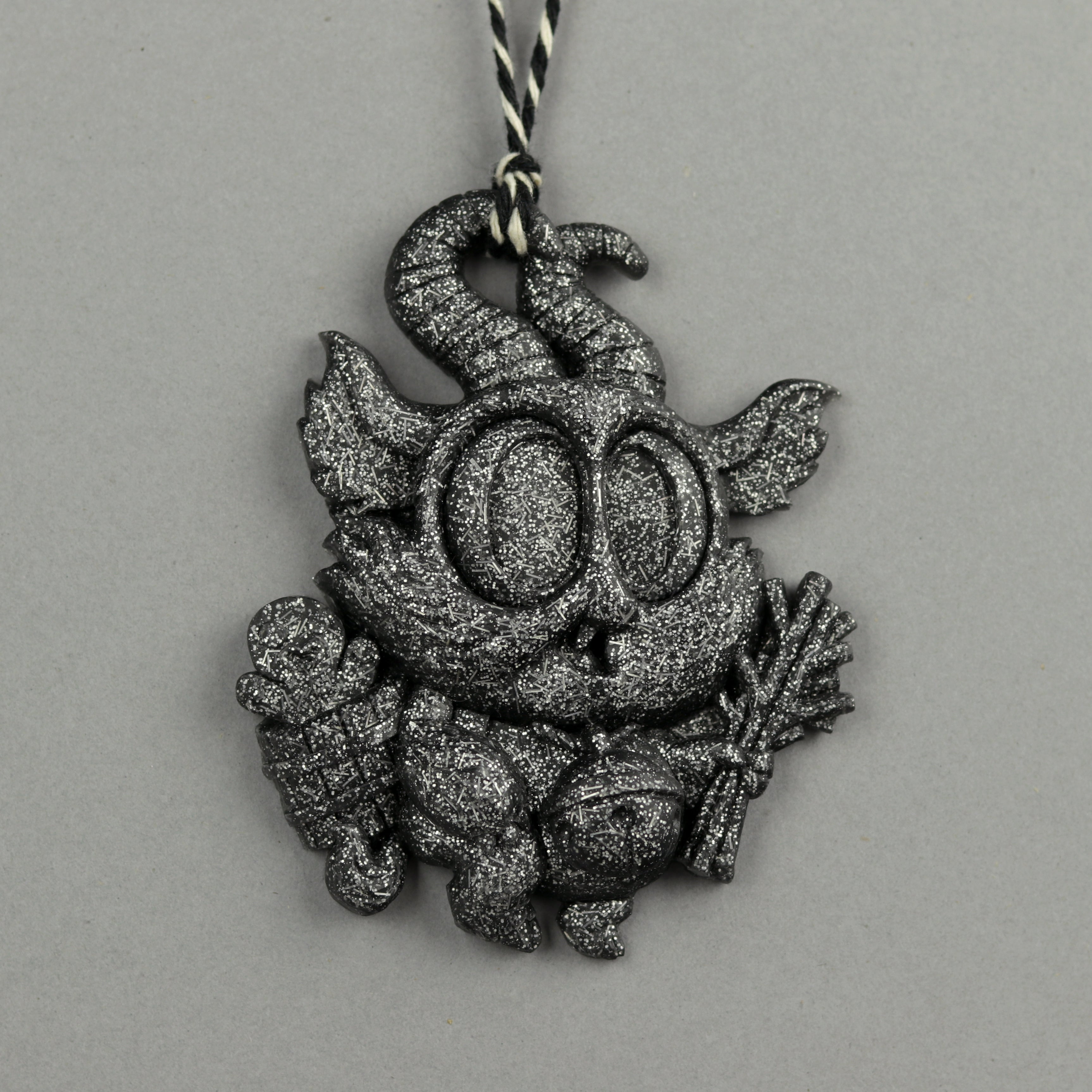 The Bindlewood Shop's Glitter Krampus Ornament features Chris Ryniak’s design in glittery black resin with big eyes, pointy ears, and two horns, holding sticks and a bag, hanging on a black-and-white string.