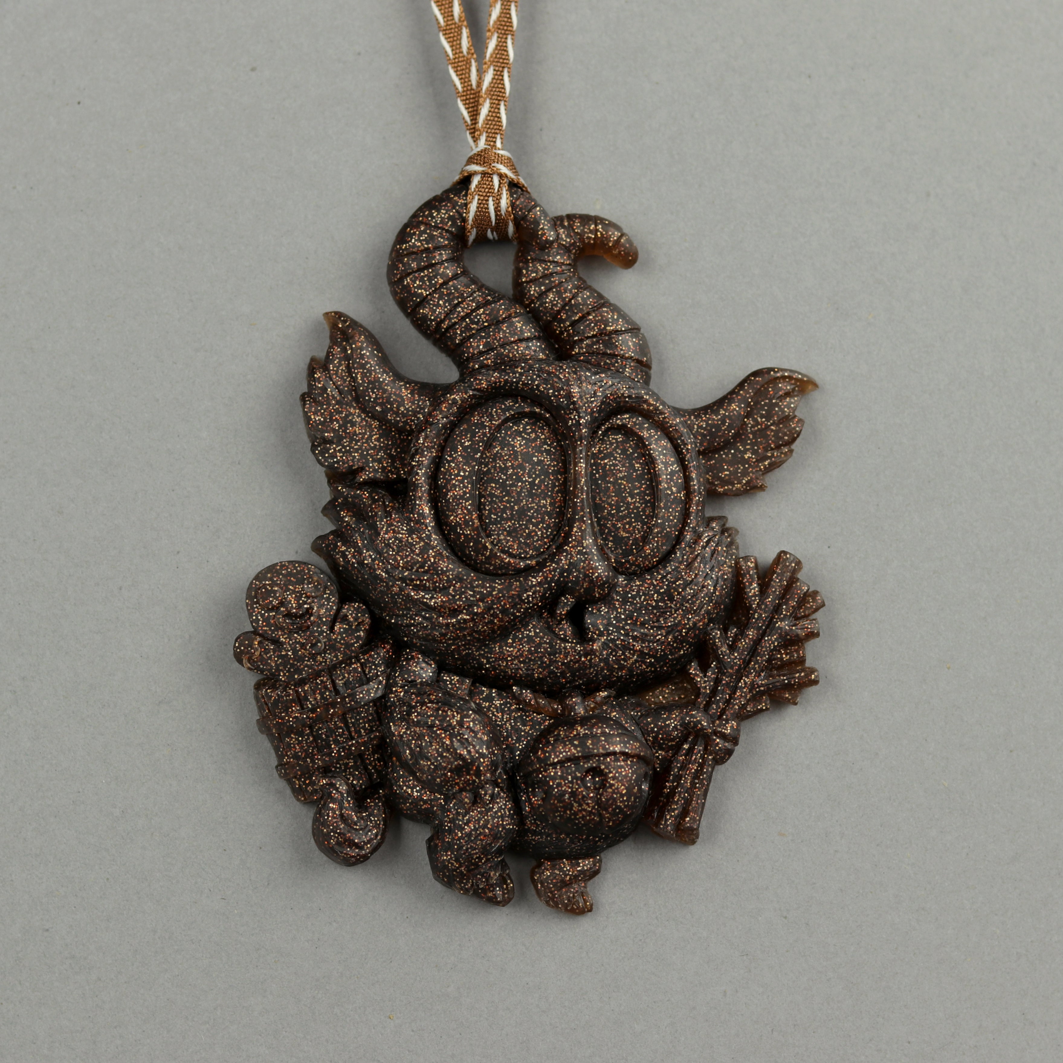 The Glitter Krampus Ornament from Bindlewood Shop features a brown, large-eyed Lil’ Krampus holding a gingerbread cookie and candy cane, hanging from a striped ribbon against a plain gray background.