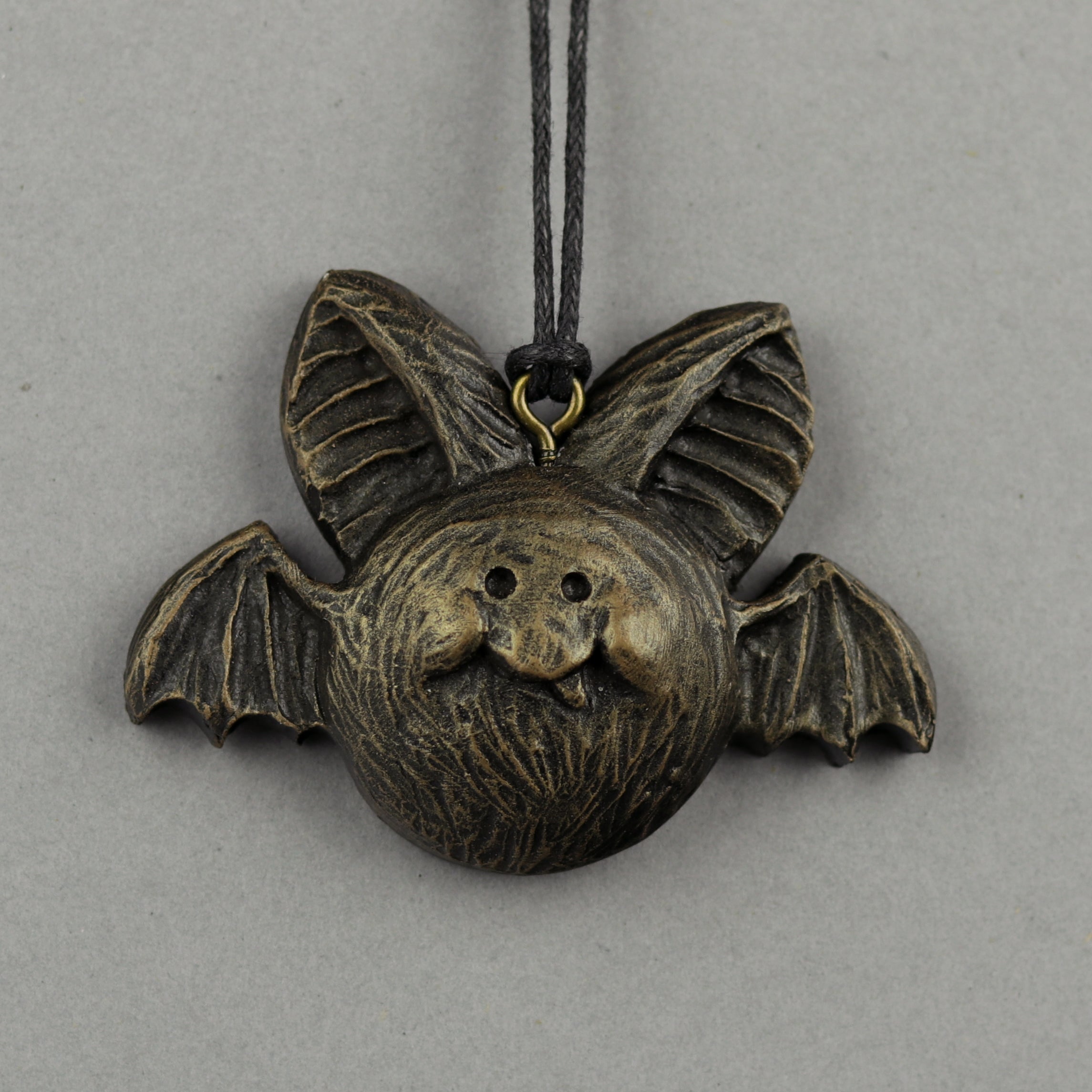 A bronze-colored Painted Mini Ornament shaped like a smiling bat with large ears and outstretched wings from Bindlewood Shop hangs on a black cord against a plain gray background.