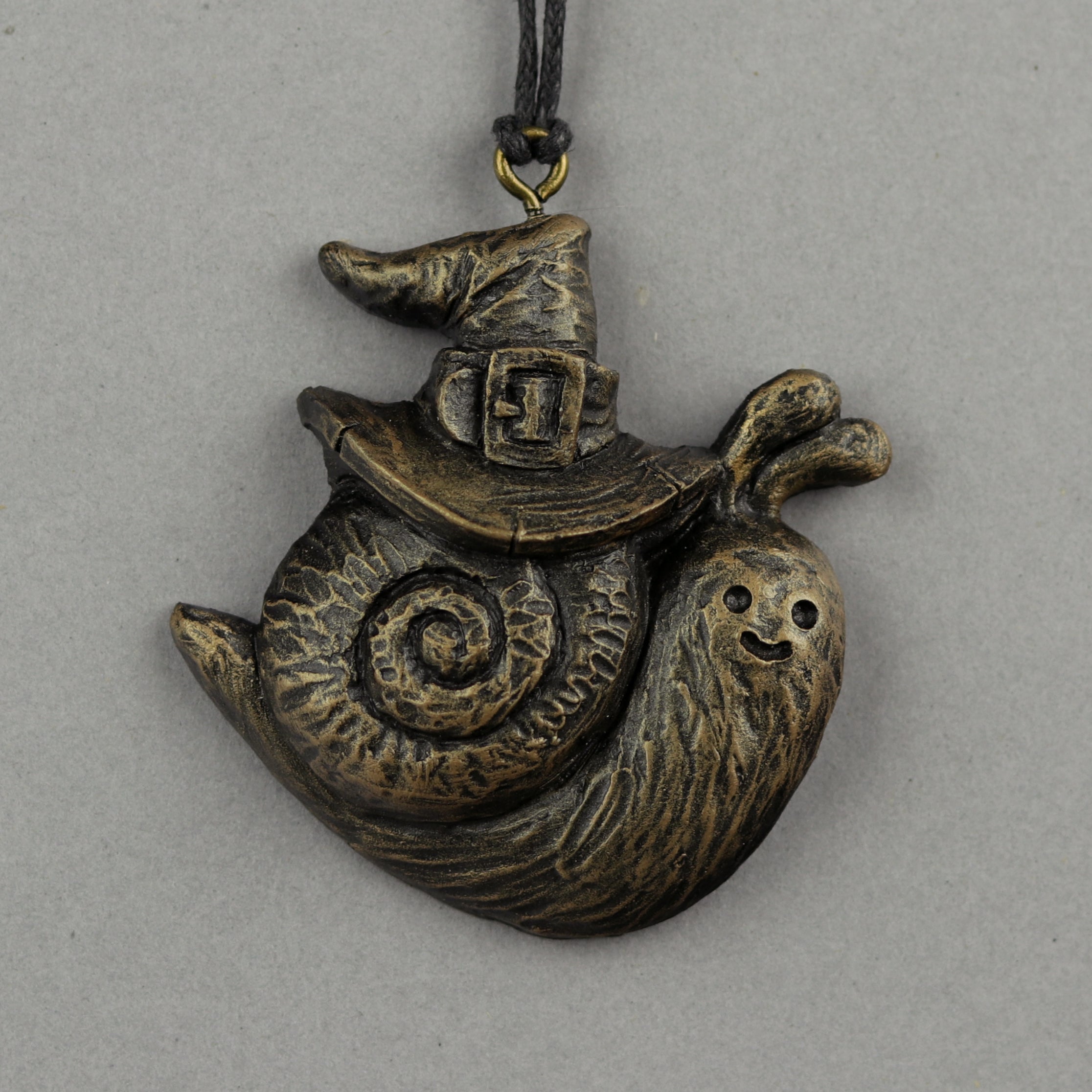A bronze-colored Painted Mini Ornament from Bindlewood Shop, shaped like a smiling snail in a pointed witch hat, inspired by Chris Ryniak’s spooky sculptures, hangs from a black cord against a gray background.