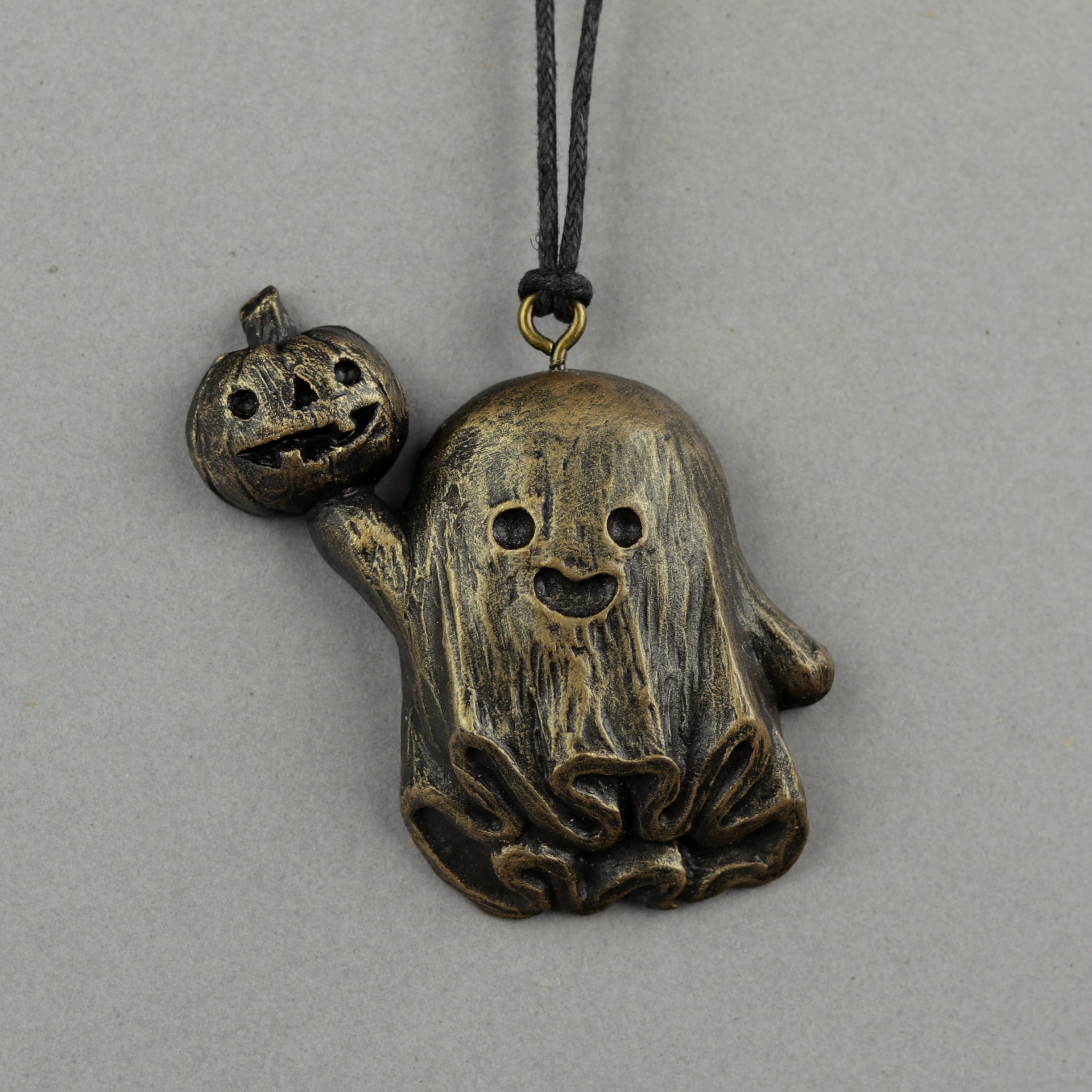 The Bindlewood Shop’s Painted Mini Ornaments feature a bronze pendant inspired by Chris Ryniak—a smiling ghost with a jack-o’-lantern—hanging from a black cord against a plain gray background.