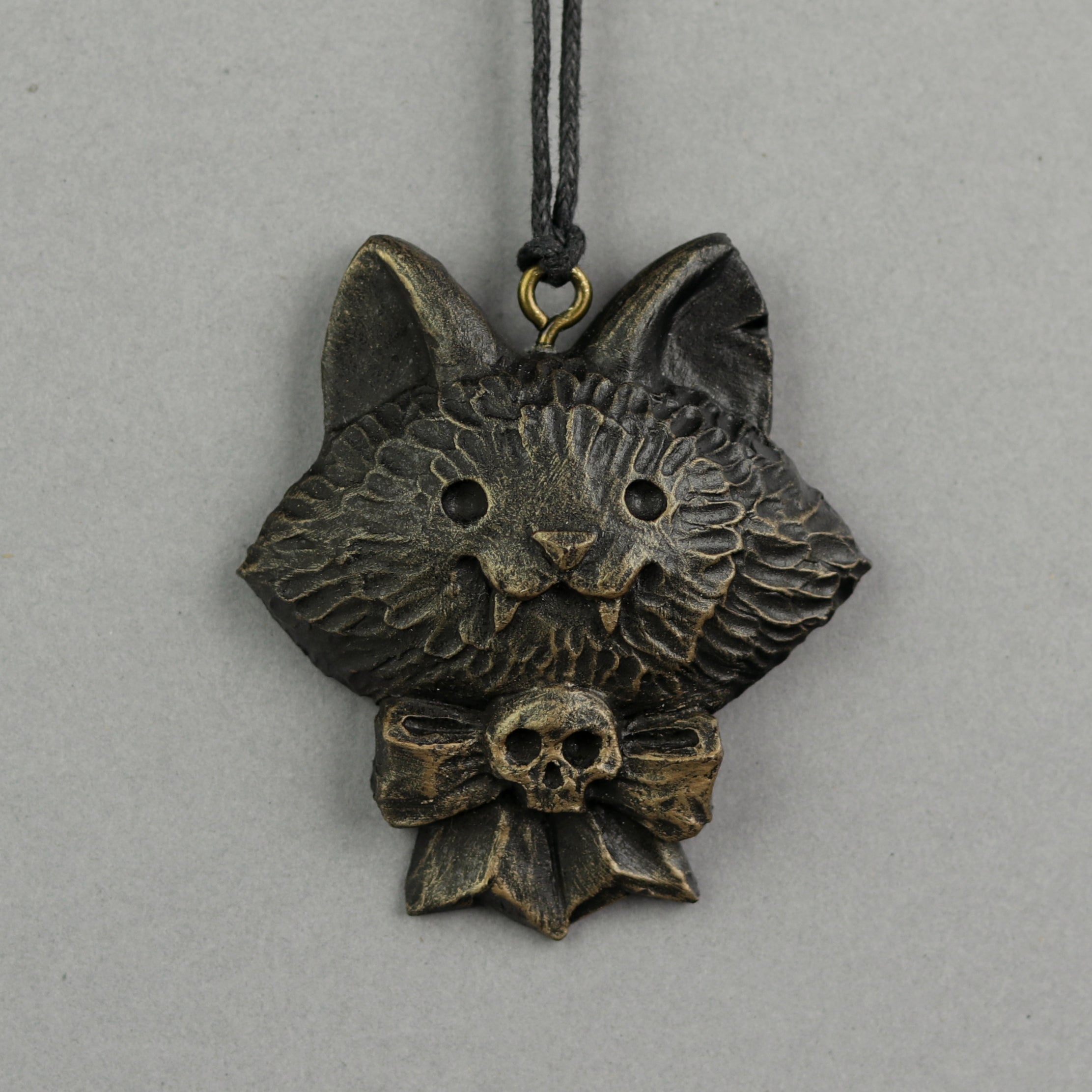 A Painted Mini Ornament from Bindlewood Shop, featuring a cat face with pointed ears, textured fur, and a skull-accented bow, hangs from a black cord against a plain gray background.