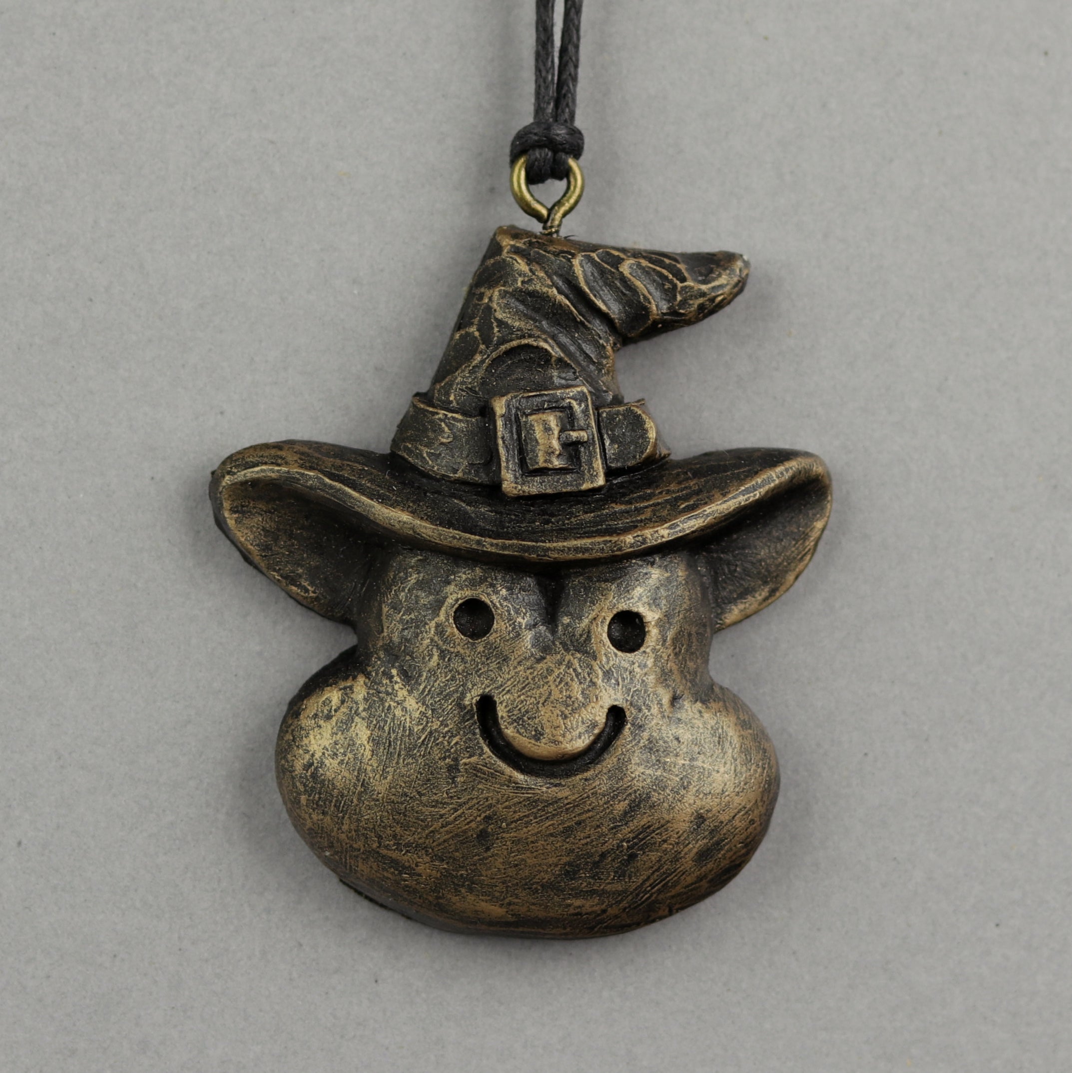 A Painted Mini Ornament from Bindlewood Shop, shaped like a smiling face with a tall, crooked witch hat and buckle, hangs from a black cord against a gray background, inspired by Chris Ryniak’s whimsical spooky sculptures.