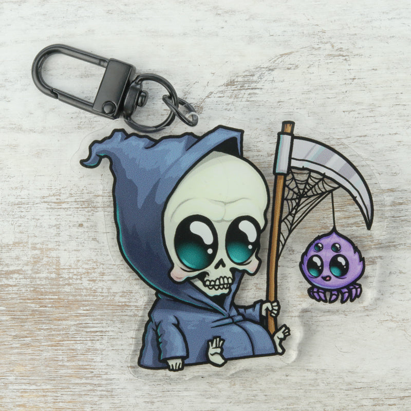 The Lil' Reaper Keychain by Bindlewood Shop features a cute Grim Reaper in a blue hood holding a scythe with a dangling purple spider, inspired by Chris Ryniak. Set against a light wooden background.