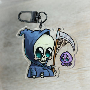 A "Lil' Reaper" acrylic charm keychain featuring a cartoon illustration of a grim reaper character in a blue cloak, holding a scythe with a cute purple spider hanging from it, inspired by Thimblestump Hollow, from Bindlewood Shop.