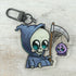 The Lil' Reaper Keychain by Bindlewood Shop features a cute Grim Reaper in a blue hood holding a scythe with a dangling purple spider, inspired by Chris Ryniak. Set against a light wooden background.