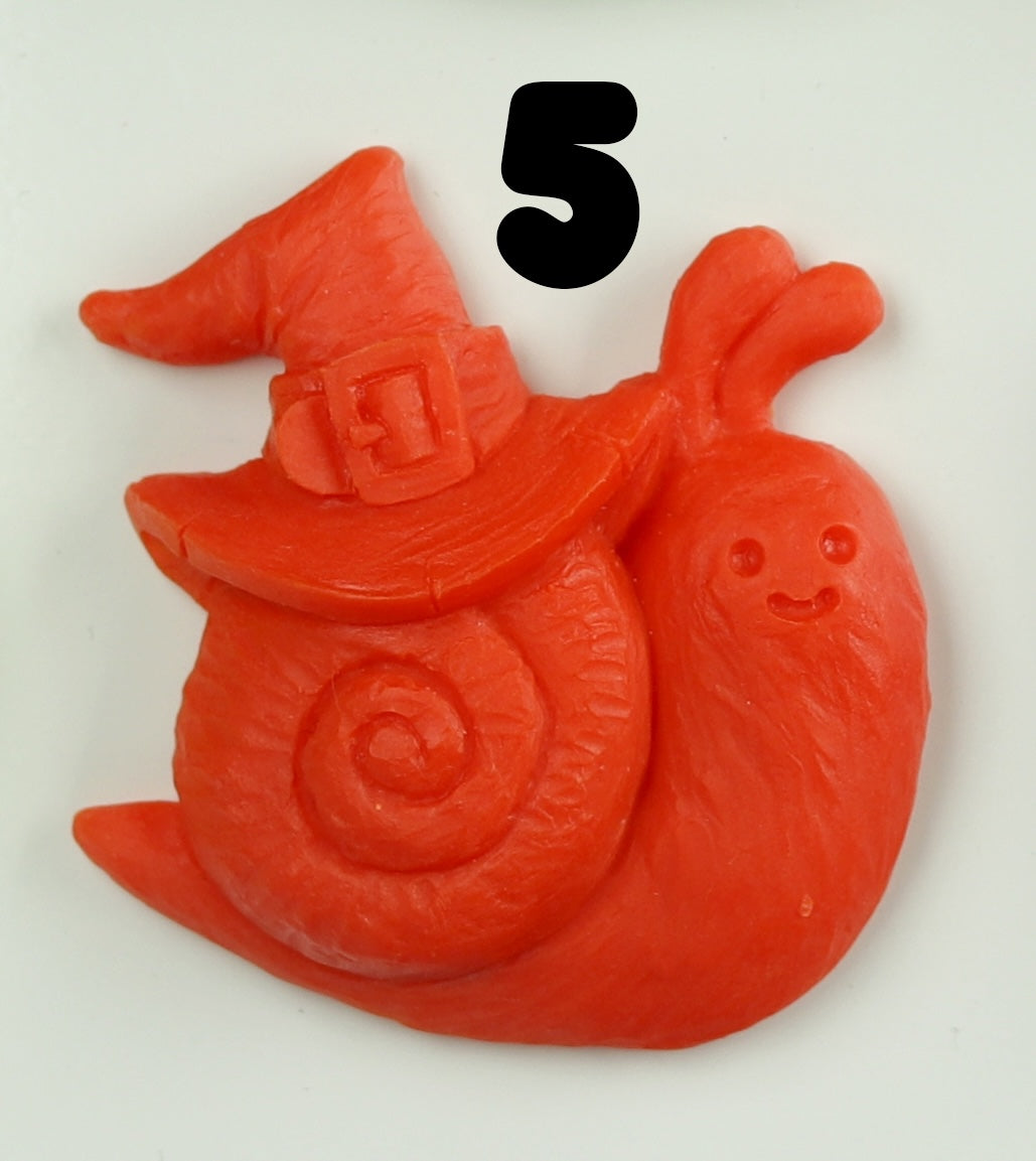 A hand-sculpted red snail magnet from Bindlewood Shop’s Halloween Magnets, featuring a smiling face, spiral shell, pointed witch hat with buckle, and a bold black “5” above its head on a white background.