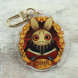A whimsical keychain featuring an illustration of a creature with rabbit ears holding a book, with ornate designs and the word "maker" printed at the bottom. This piece is part of "The Maker" Acrylic Charm by Bindlewood Shop.