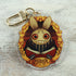 The "The Maker" Keychain by Bindlewood Shop features an acrylic charm of a wide-eyed, grinning creature with rabbit ears, framed by golden leaves and holding an open red book labeled "The Maker.