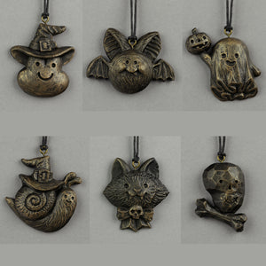 Six painted mini ornaments by Bindlewood Shop—bronze Halloween charms inspired by Chris Ryniak: a smiling witch hat, bat, ghost with pumpkin, snail witch, cat with bow/skull, and skull with crossbones—all on black cords.