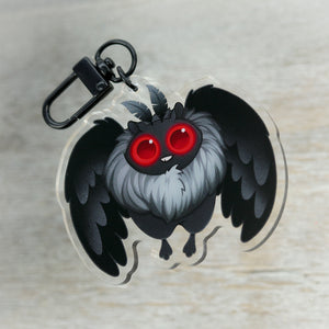 An "Mothman" acrylic charm featuring a stylized, cartoonish black owl from Thimblestump Hollow with striking red eyes and a fluffy grey chest, perched and looking intently forward by Bindlewood Shop.