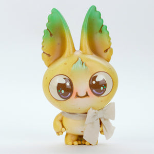 A cute, round-eyed creature called Yellow Kangorumple, from Bindlewood Shop, stands against a plain white background. This hand-painted figure, featuring large green-tipped ears and a gradient yellow-green body, has a small smile and holds a white ribbon in its left hand. It resembles the charming style of Chris Ryniak’s special edition collectibles.