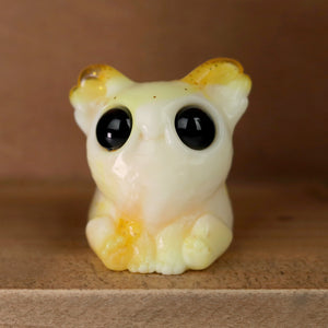 A small, cute ceramic creature with large black eyes and tiny arms is sitting on a wooden surface. Reminiscent of Chris Ryniak's whimsical designs, the creamy yellow figurine from the 3D Matinee Set, with slight orange accents on its ears and feet, exudes an endearing charm akin to resin figures like Smidgen and Dashi.