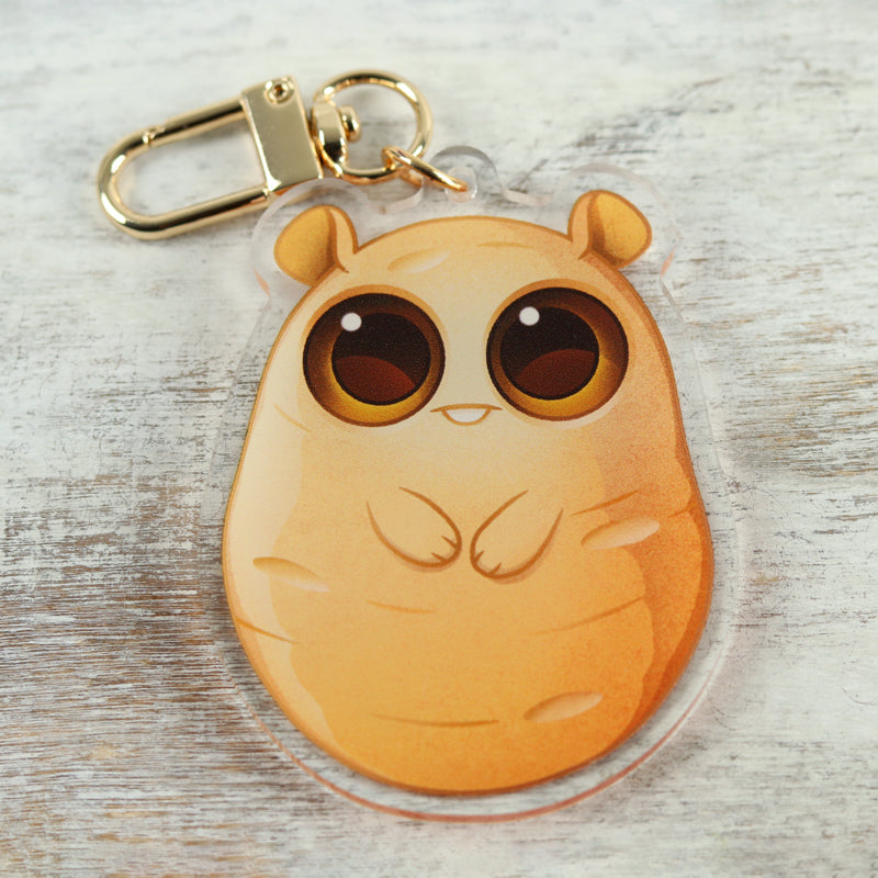 The Lucky Potato Keychain by Bindlewood Shop is a cute acrylic charm featuring a chubby, cartoon-style orange creature with big round eyes—resembling a potato—lying on a light wooden surface. Inspired by Amanda Louise Spayd's whimsical style.