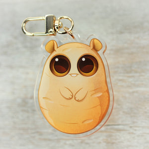 A cute, cartoon-style "Lucky Potato" Acrylic Charm keychain designed by Chris Ryniak or Amanda Spayd for Bindlewood Shop, with big, expressive eyes and a chubby appearance, set against a wooden.