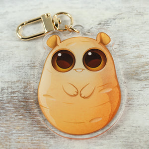 Cartoon-style "Lucky Potato" Acrylic Charm keychain with large, soulful eyes designed by Chris Ryniak, on a rustic wooden surface. Brand: Bindlewood Shop.
