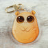 The Lucky Potato Keychain by Bindlewood Shop is a cute acrylic charm featuring a chubby, cartoon-style orange creature with big round eyes—resembling a potato—lying on a light wooden surface. Inspired by Amanda Louise Spayd's whimsical style.