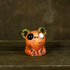 A small, orange Rotten Pumpkin Smidgen by Chris Ryniak, featuring a koala-like shape with one large black eye and one large white eye, sits on a wooden surface against a blurred brown background. This adorable handmade and hand-painted resin figurine showcases the whimsical creature design style Chris Ryniak is known for.